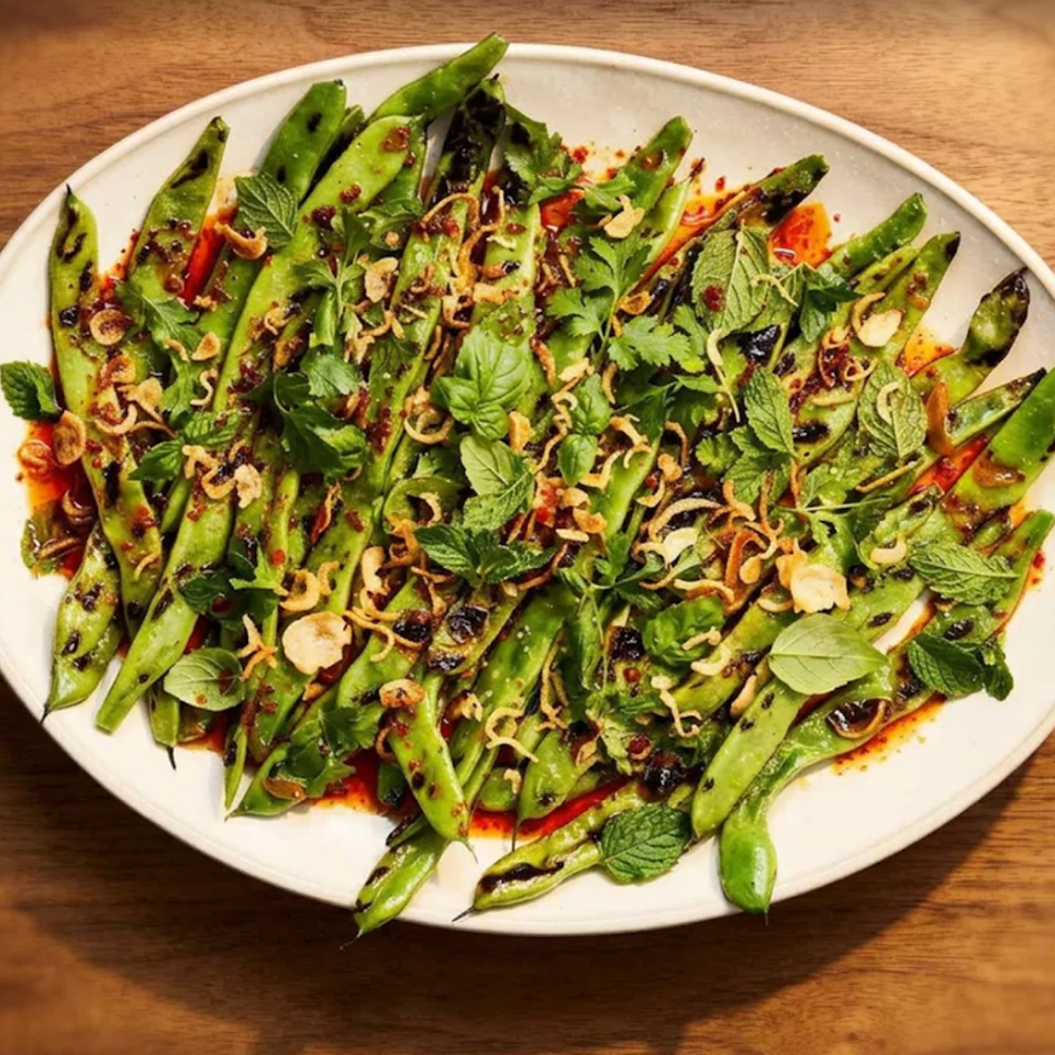 Grilled green beans and snap peas topped with fresh herbs, crispy shallots, and chili oil on a white platter.