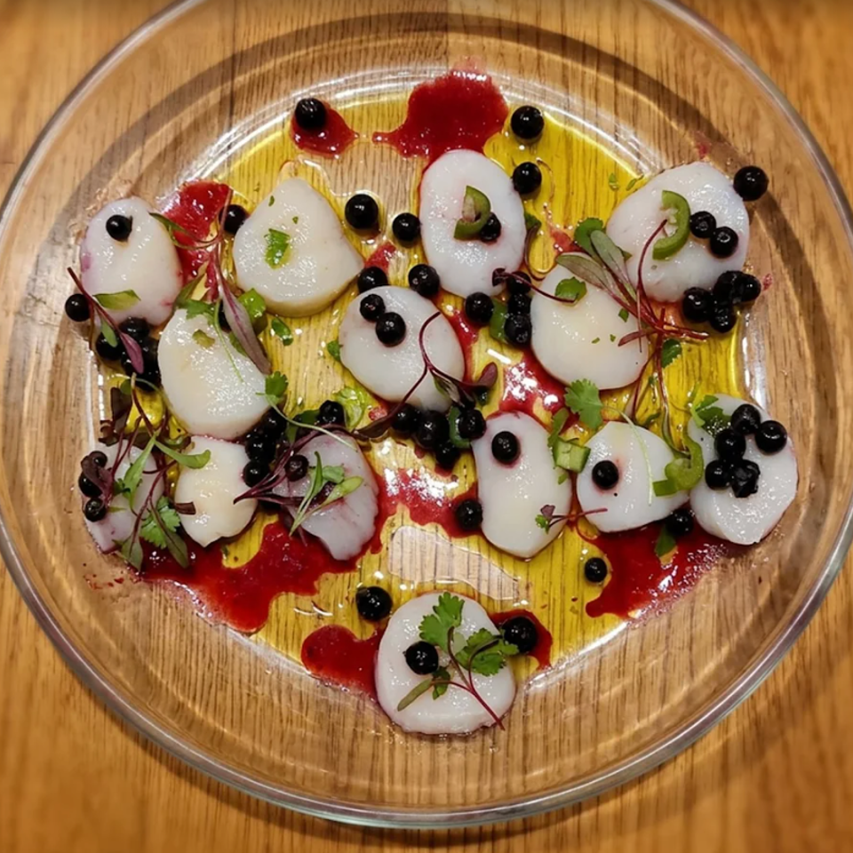 Overhead view of scallops arranged with blackberries, microgreens, olive oil, and red berry sauce on a glass plate.